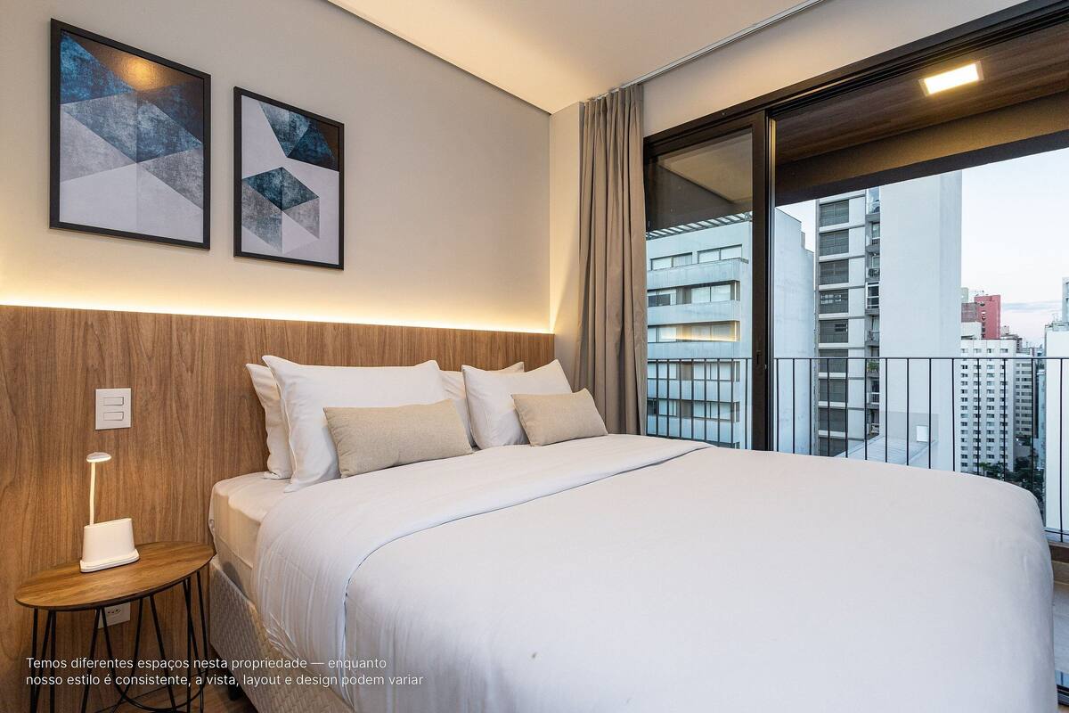 Une chambre d’hôtel avec un grand lit, une table de chevet, deux œuvres d’art encadrées et une vue sur la ville par une grande fenêtre.