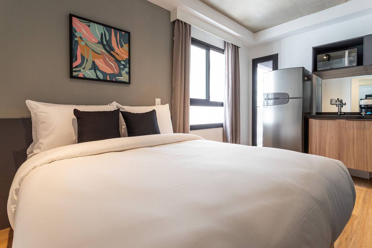 Une chambre d’hôtel avec un lit, un tableau au mur, une fenêtre avec des rideaux, un coin cuisine avec un réfrigérateur et un four à micro-ondes.