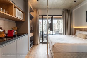 A modern hotel room with a bed, a kitchen area, and a balcony with a city view.
