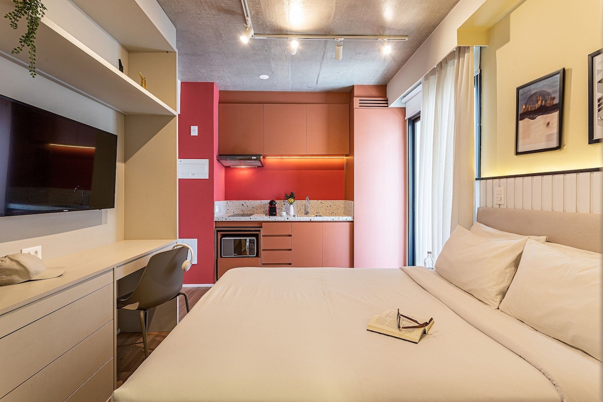 A hotel room with a bed, desk, chair, kitchenette, and a TV.
