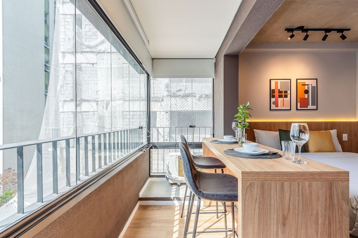 Une chambre d’hôtel moderne avec un balcon, une table à manger en bois, deux chaises, un lit et deux œuvres d’art encadrées accrochées au mur.