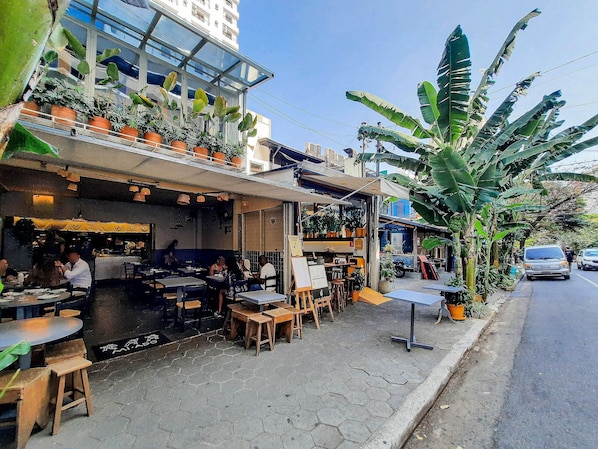 An outdoor cafe with tables and chairs, surrounded by potted plants and a building in the background.