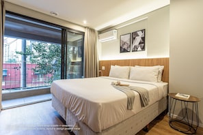 A modern hotel room with a large bed, a small round table, and a view of the city through a large window.