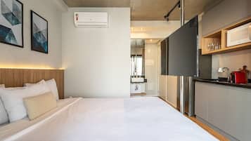 Quarto de hotel moderno com uma cama grande, uma cozinha compacta e um banheiro visível ao fundo.