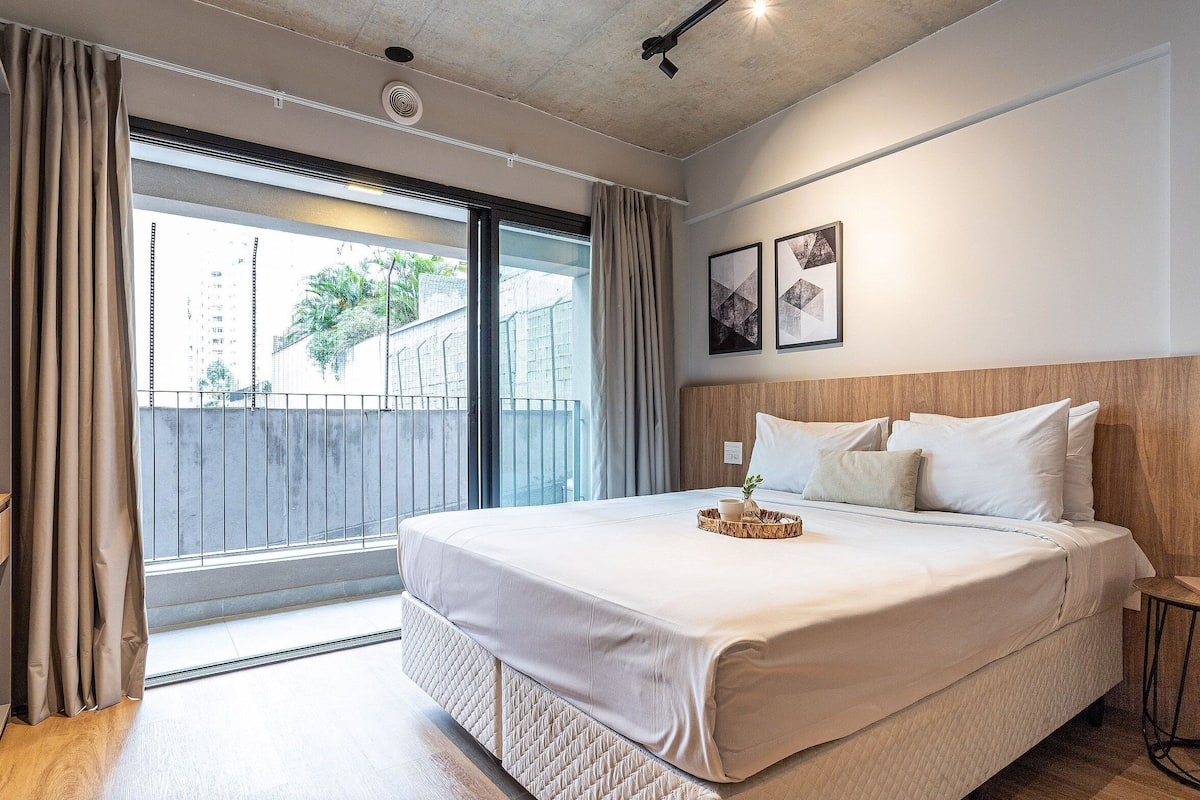 Une chambre d’hôtel avec un grand lit, une petite table de chevet, une fenêtre avec des rideaux et une vue sur les bâtiments environnants.
