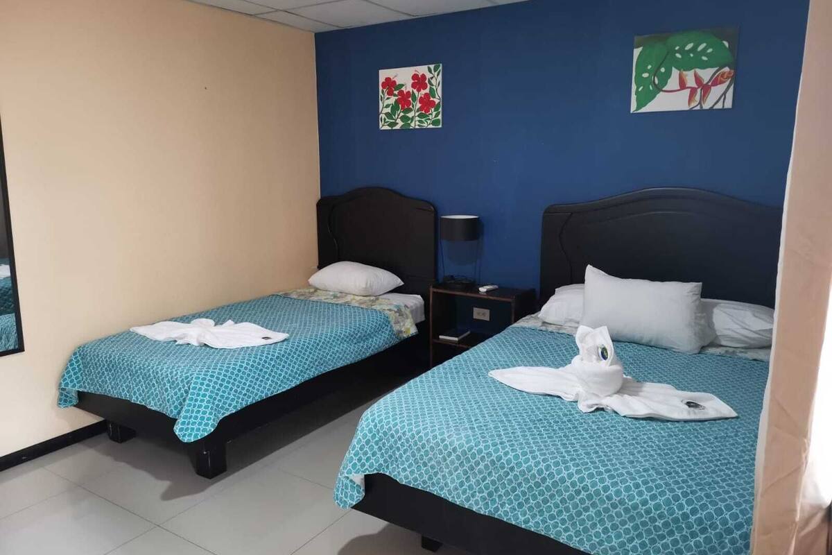 A hotel room with two single beds, each with a blue patterned bedspread and white pillows. There are two wall-mounted paintings, one with red flowers and another with green leaves.