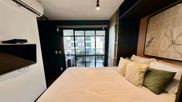 A modern hotel room with a large bed, a flat-screen TV, and a view of the city through large windows. - Ator - Chic Studio in Vila Olimpia (São Paulo)