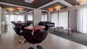 A modern hotel lobby with a round red table, dark purple chairs, and a sofa. There are large windows with white curtains and a ceiling with recessed lighting. - Ator - Chic Studio in Vila Olimpia (São Paulo)