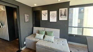 A modern living room with a grey sofa, green and white patterned pillows, and framed artwork on the wall. - Ator - Chic Studio in Vila Olimpia (São Paulo)