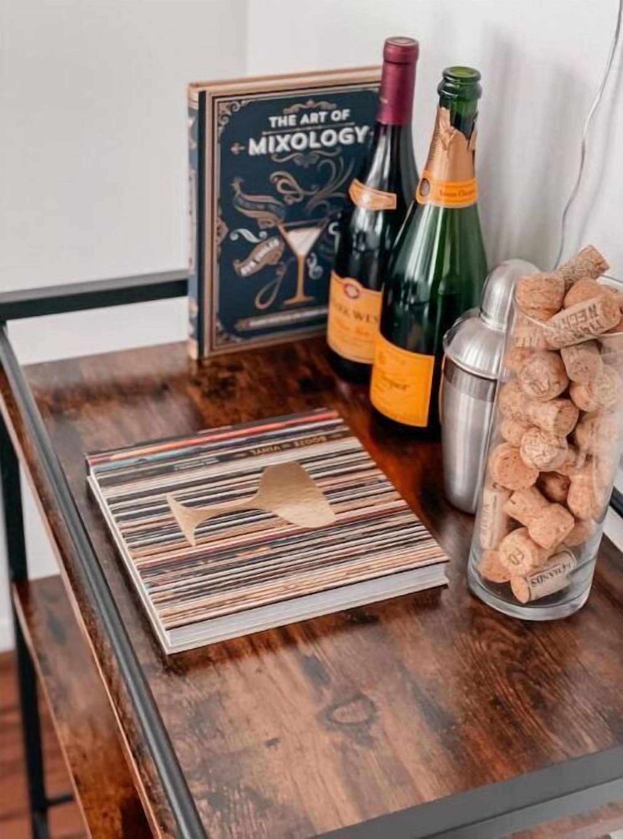 A wooden table with a framed book titled "The Art of Mixology," two bottles of wine, a cocktail shaker, and a jar of corks.