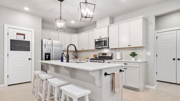 Una cocina moderna con armarios blancos, una isla central con taburetes, electrodomésticos de acero inoxidable e iluminación colgante.