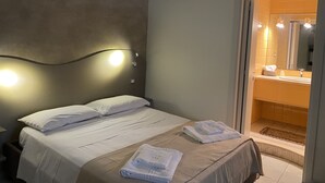 Une chambre d’hôtel avec un lit, une table de chevet, une lampe fixée au mur et une salle de bain visible à travers une porte ouverte.