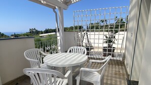 Une terrasse couverte avec une table ronde et quatre chaises en plastique blanc, une clôture en métal et des plantes en pot.