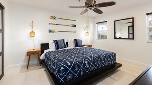 A bedroom with a bed, two nightstands, a ceiling fan, a wall-mounted mirror, and a window with a view of the outdoors.