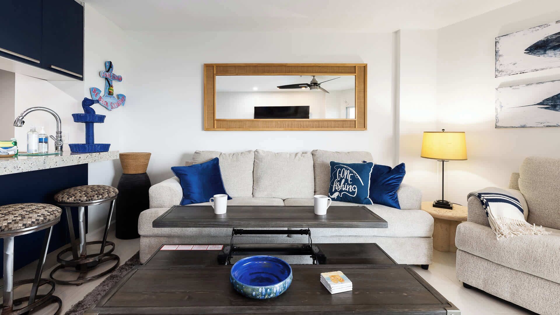 A living room with a sofa, coffee table, and a TV. There is a kitchen area with a sink and a mirror.