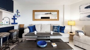 Una sala de estar con sofá, una mesa de centro y televisión. Hay una zona de cocina con un lavamanos y un espejo.
