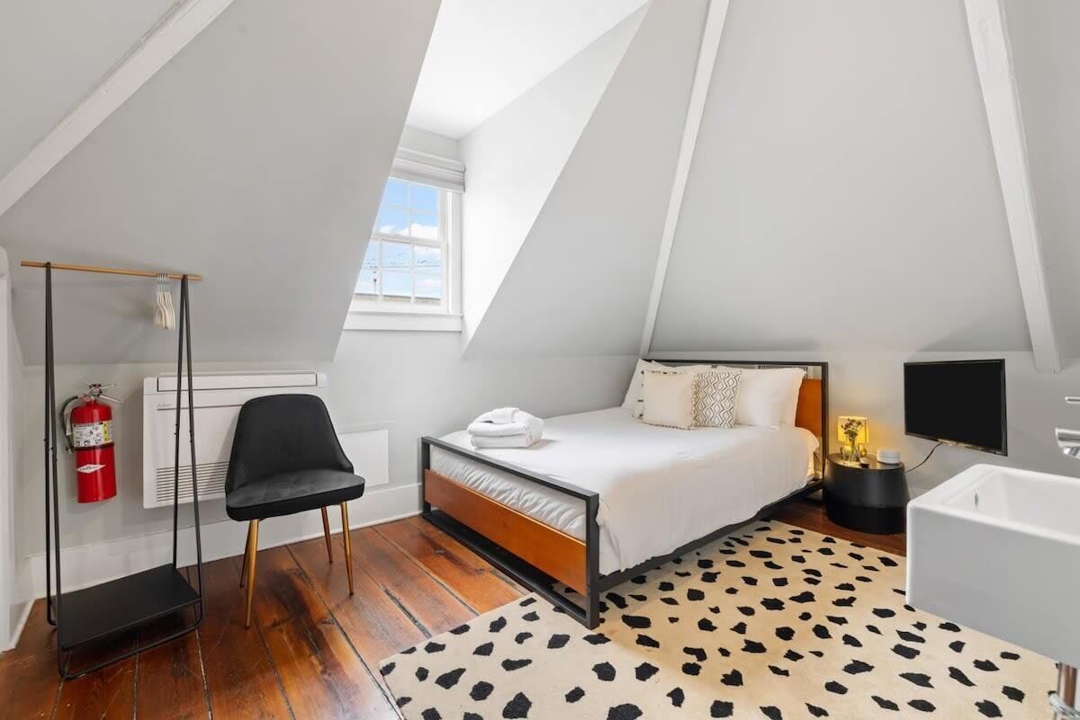 A bedroom with a sloped ceiling, a bed, a chair, a fire extinguisher, a TV, and a bathroom with a bathtub.