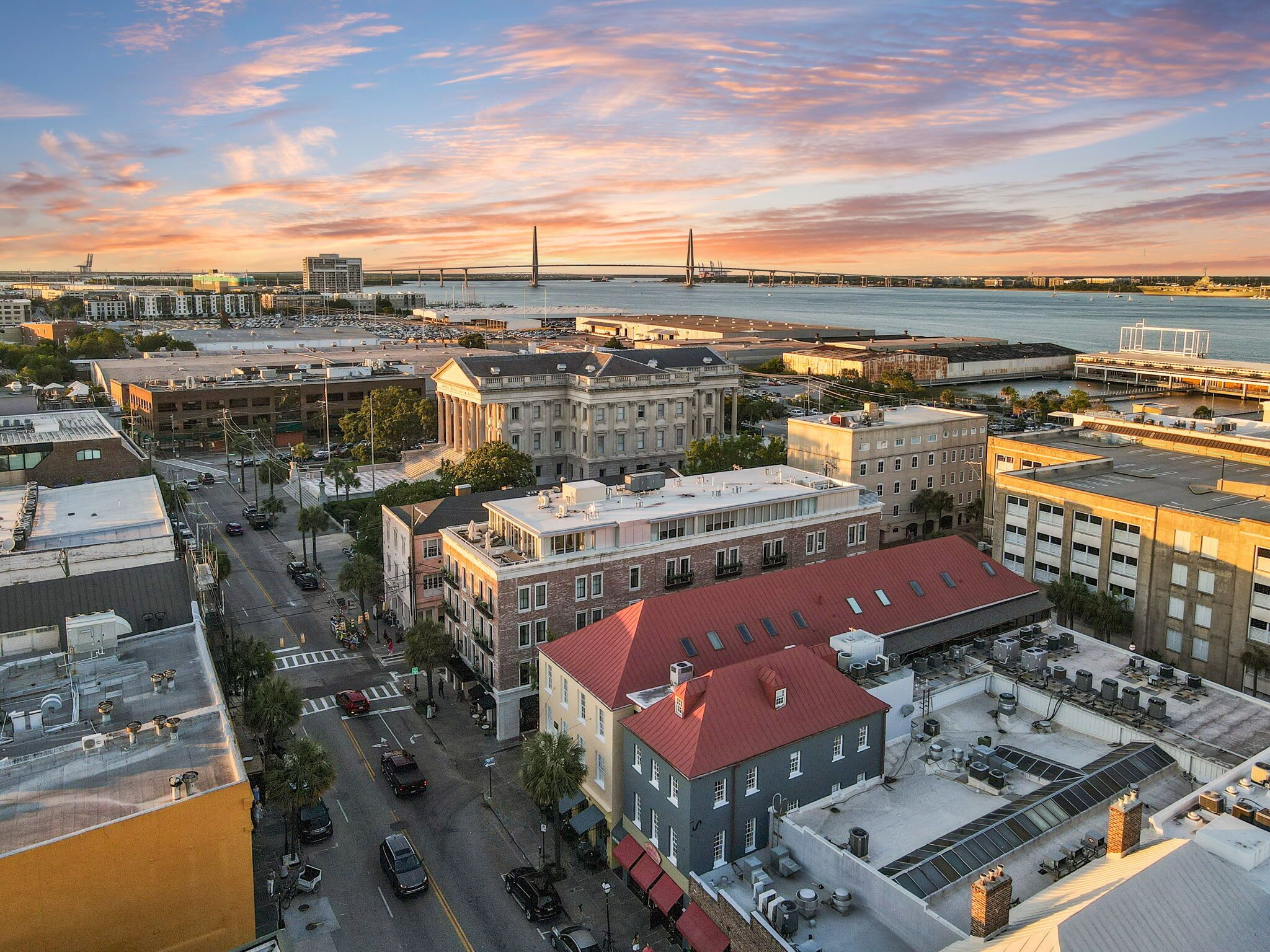 An aerial view of a cityscape with a prominent building featuring a red roof and multiple windows, surrounded by other buildings and a bridge in the background.
