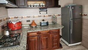 Een keuken met donkere houten kastjes, een gasfornuis, een rode pan, een koelkast en een betegelde achterwand.
