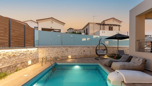 Una piscina con un sillón de tomar el sol y una silla colgante, rodeada de muros de piedra y una cerca de madera.