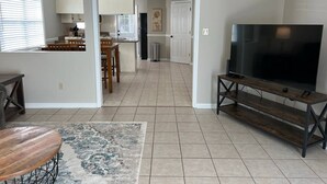 A spacious living area with a large TV, a dining table, and a kitchen in the background. - Get-Away on Wayland Cozy Cottage (Valdosta)