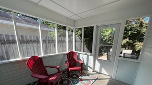 A sunroom with two red Adirondack chairs, a patterned rug, and a view of a wooden fence and trees. - Get-Away on Wayland Cozy Cottage (Valdosta)