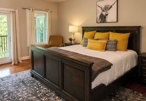Une chambre à coucher avec un grand lit, une chaise, une table de chevet, une lampe, une fenêtre avec des rideaux et un tableau encadré au mur.