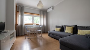 Un salon moderne avec un canapé gris, une table à manger avec des chaises, un téléviseur et une grande fenêtre avec des rideaux.