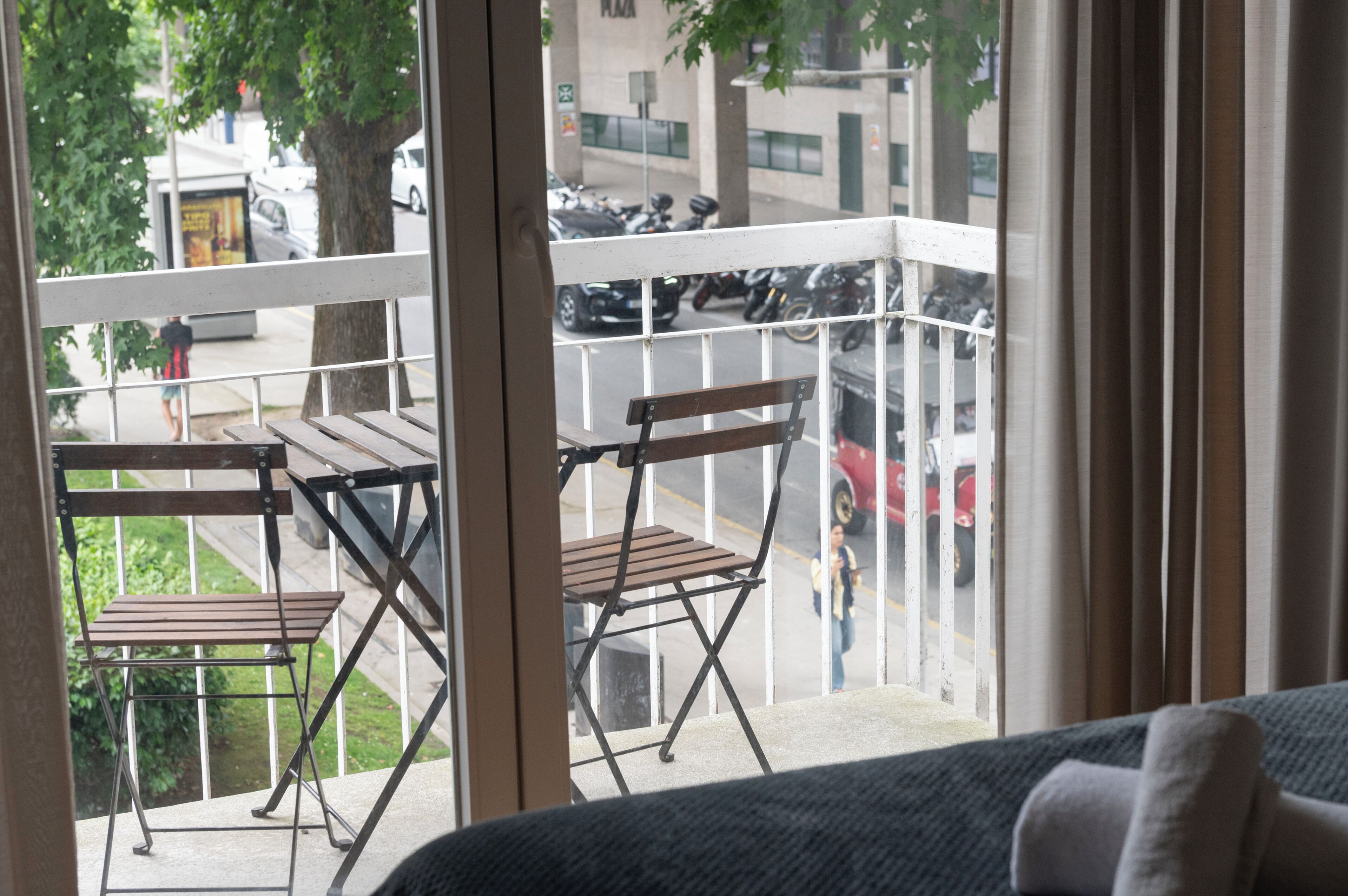 A balcony with a wooden table and two folding chairs, overlooking a street with parked cars and a pedestrian.