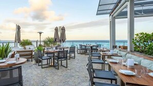 Ein Sitzbereich im Freien mit Tischen, Stühlen, einer Couch und Sonnenschirmen mit Blick auf das Meer.