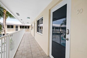 Um corredor com uma porta identificada com o número '30', uma varanda com piscina e uma palmeira.