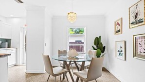 Un espace repas moderne avec une table en verre, quatre chaises, un luminaire suspendu et des œuvres d’art encadrées accrochées au mur.