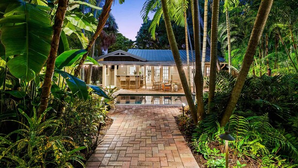 Une maison avec un chemin de briques menant à un porche vitré, entourée d’une végétation tropicale luxuriante.
