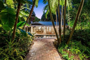 A house with a brick pathway leading to a glass-enclosed porch, surrounded by lush tropical vegetation.
