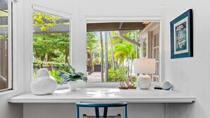 Un bureau blanc donnant sur un jardin luxuriant, une chaise bleue et un tableau encadré accroché au mur.