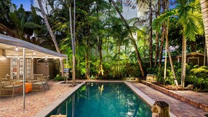 Une piscine rectangulaire entourée d’une végétation luxuriante et un patio couvert aménagé avec des meubles en osier.