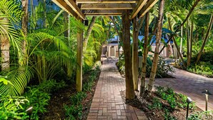 Une allée couverte, avec des poutres en bois, bordée d’une végétation tropicale luxuriante et d’un chemin en briques.