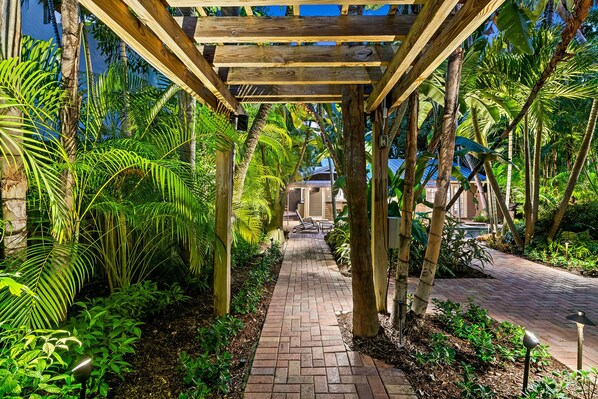 A covered walkway with wooden beams, surrounded by lush tropical plants and a brick pathway.