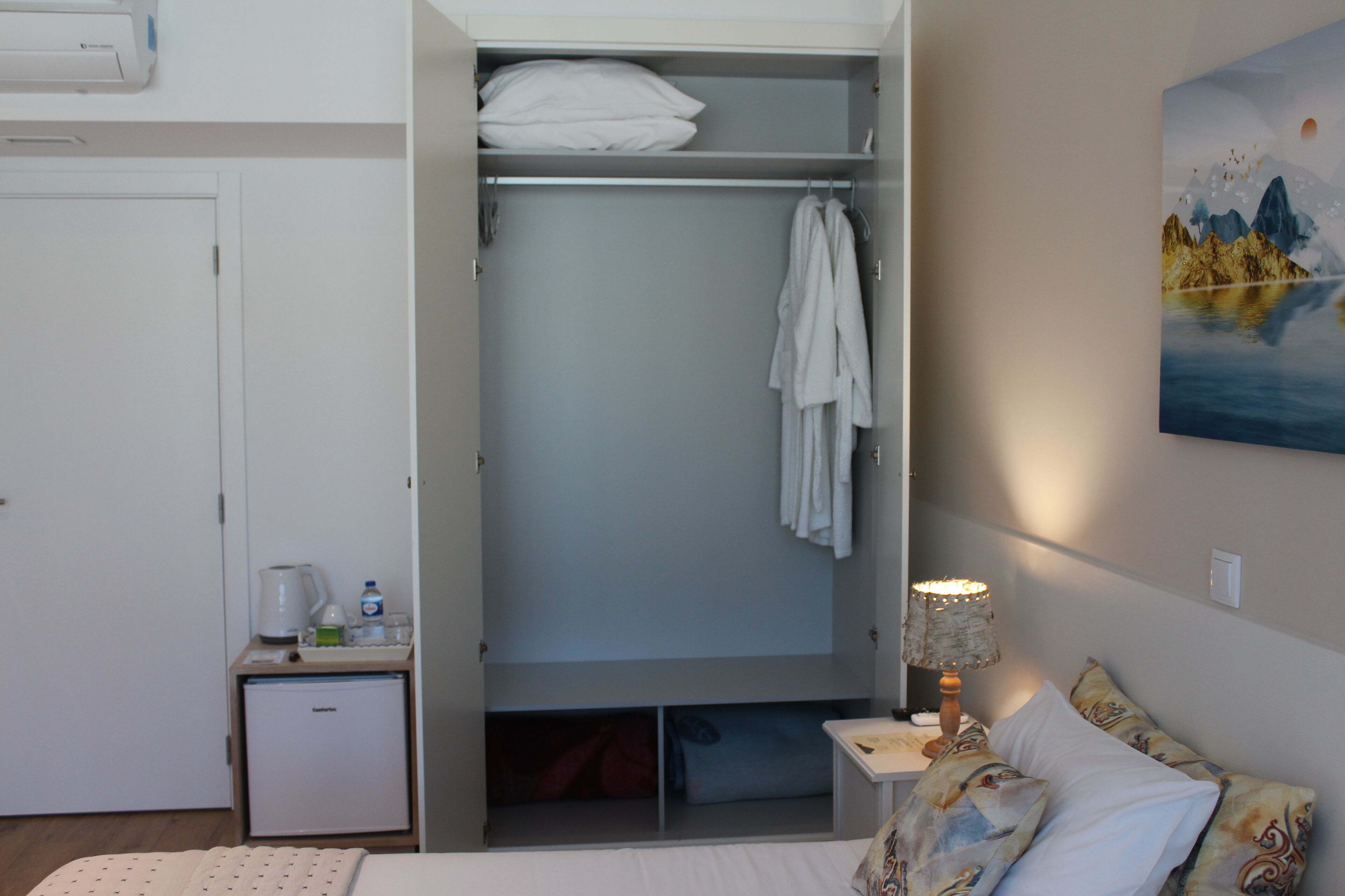 A bedroom with a bed, a bedside table with a lamp, a small bedside table with a kettle and glasses, a wardrobe, and a painting on the wall.