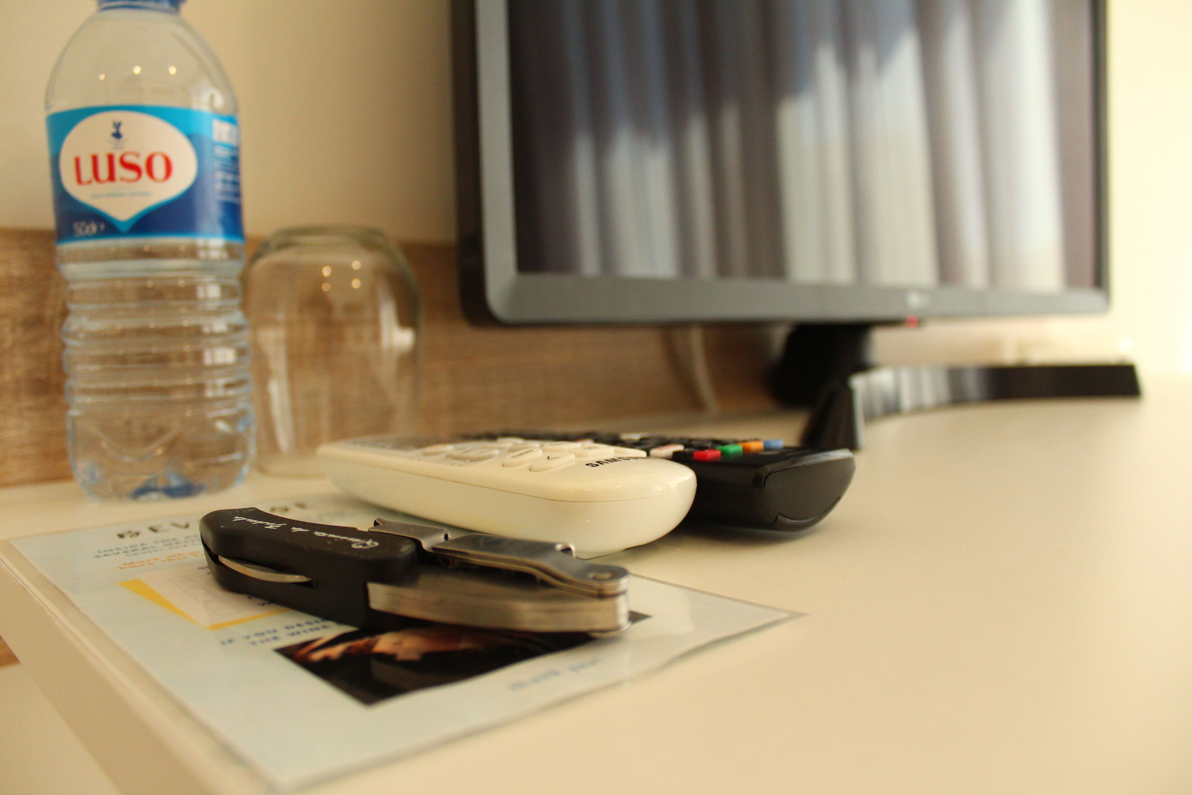 A hotel room with a desk, a bottle of Luso water, a remote control, and a keychain with a photo.