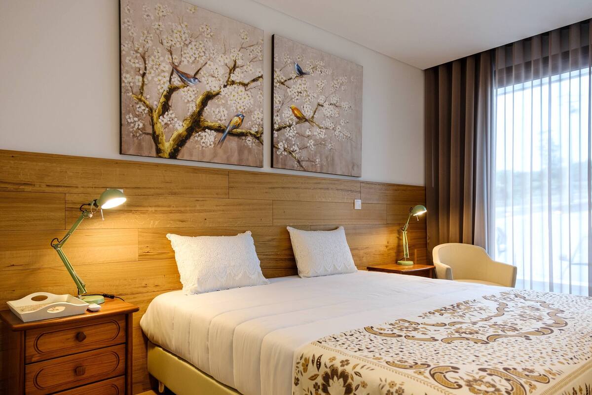 Habitació d'hotel amb un capçal de llit de fusta, un llit gran amb roba de llit blanca, dos quadres penjats a la paret amb representació d'ocells, una finestra amb cortines i una tauleta de nit amb una làmpada.