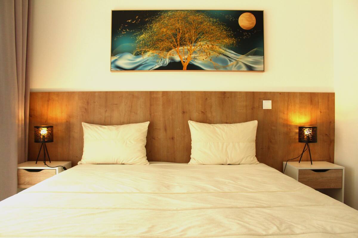 Habitació d'hotel amb un capçal de llit de fusta, dues taules de nit amb làmpades, un llit amb roba de llit blanca i un quadre amb un arbre i una lluna a la paret.
