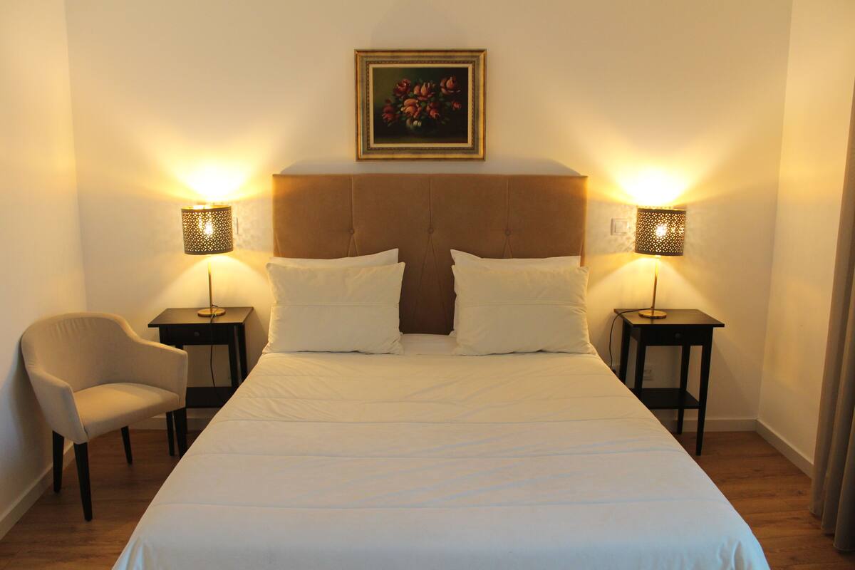 Una habitació d'hotel amb un llit gran, dues taules de nit amb làmpades, una cadira i un quadre a la paret.
