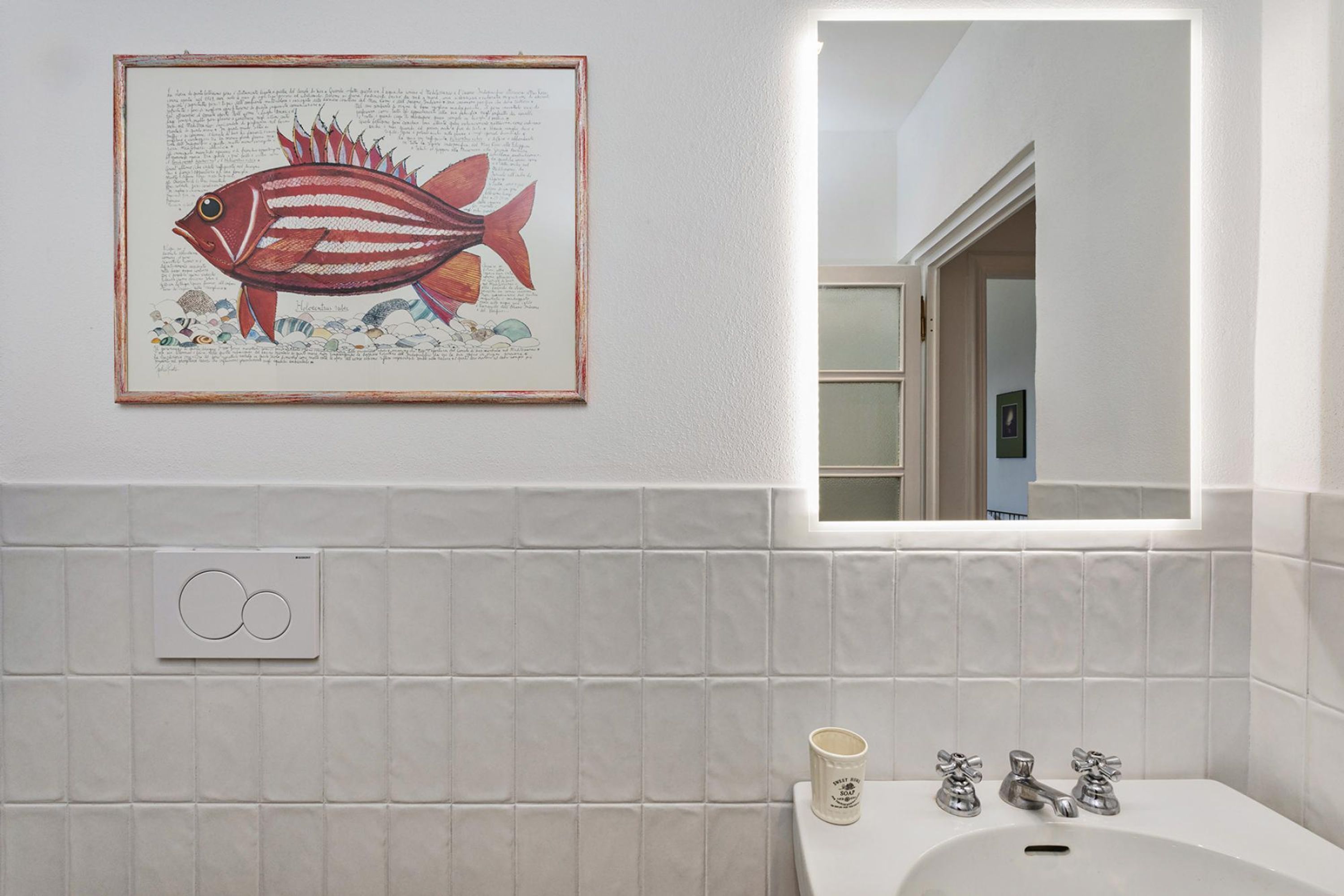 A bathroom with white tiles, a framed fish illustration, a mirror, and a sink with two faucets.
