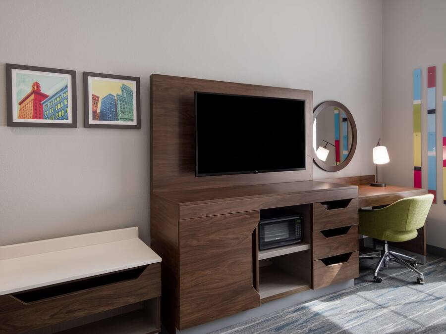 A modern hotel room with a wooden entertainment unit, a flat-screen TV, a desk, a green office chair, and two framed cityscape pictures on the wall.
