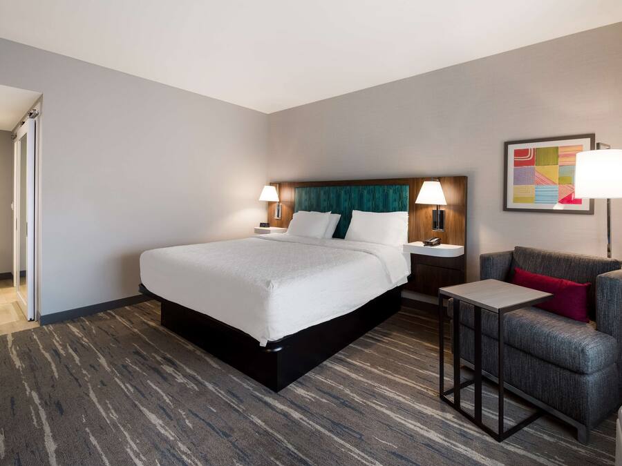 A hotel room with a large bed, a small table, a chair, and a nightstand with a lamp.