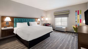 A hotel room with a large bed, bedside tables with lamps, a desk with a chair, a window with a view, and a colorful artwork on the wall.