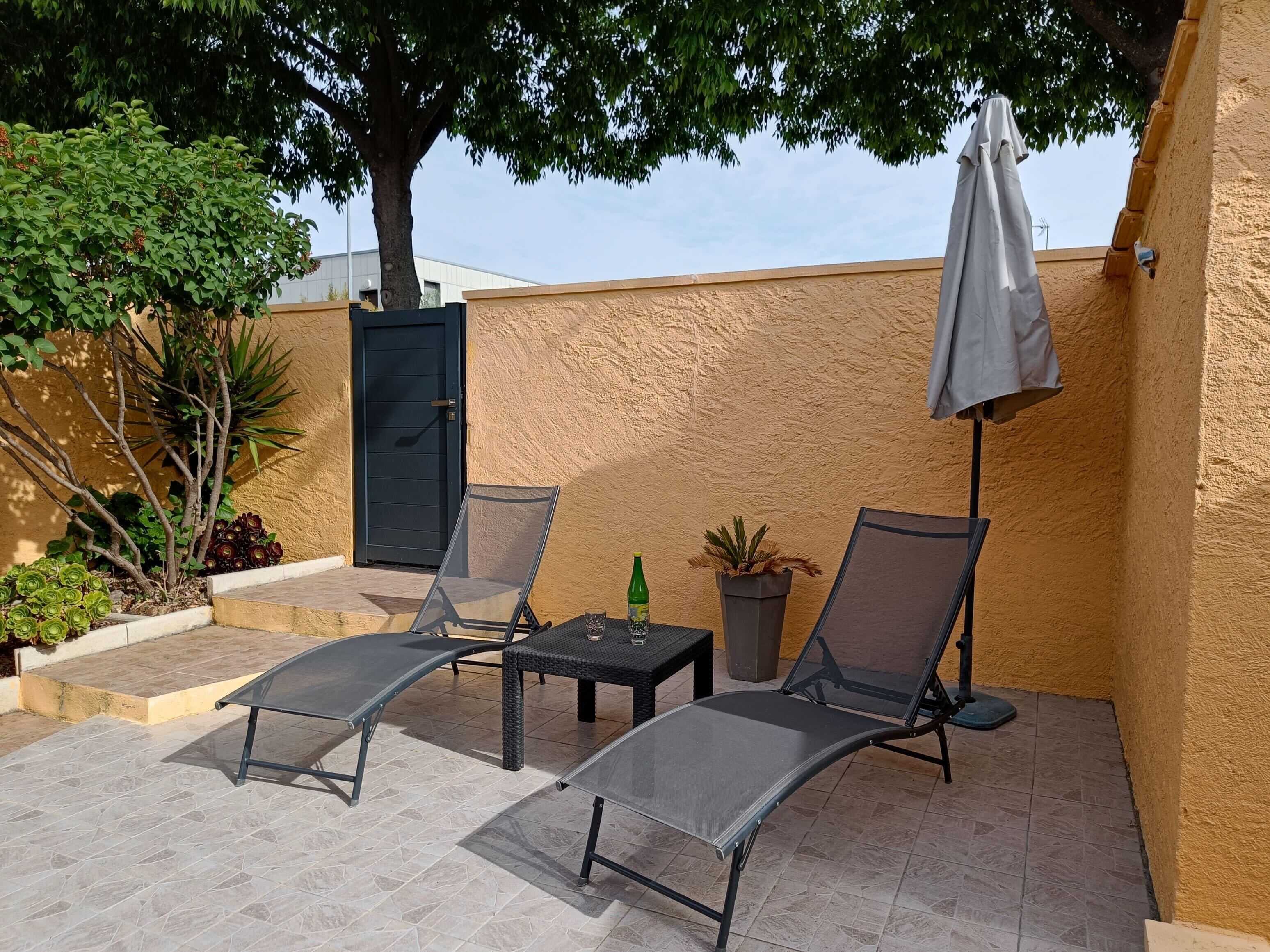 An outdoor patio with two reclining chairs, a small table, a potted plant, and a door.