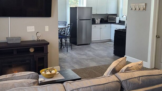 A modern living room with a sofa, a coffee table, and a TV. A kitchen with white cabinets and a refrigerator is visible in the background. - Location, Location in cedarburg (Cedarburg)