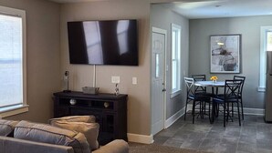 A living room with a flat-screen TV, a dark entertainment unit, and a sofa. There is a dining area with a table and chairs, and a kitchen area with a refrigerator. - Location, Location in cedarburg (Cedarburg)